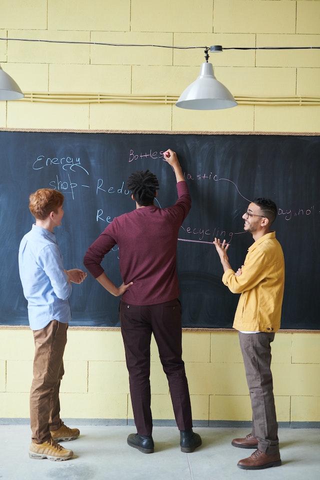 Group of men standing at a black board solving problems.